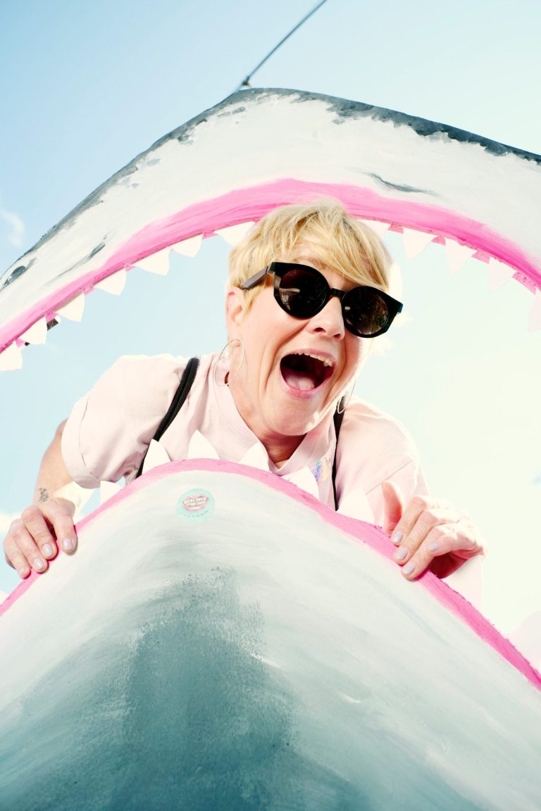 Frau mit Sonnenbrille, die begeistert aus dem Maul eines weißen Haifischs schaut.
