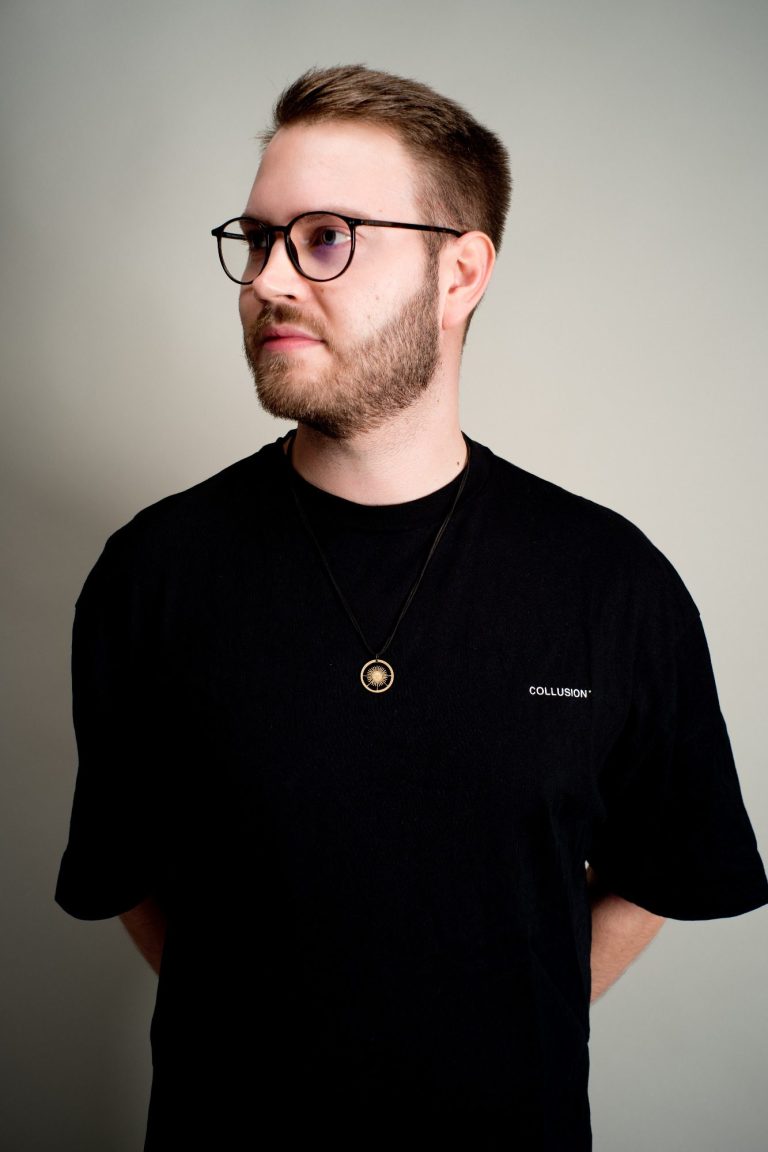 Mann mit Brille, schwarzem Shirt und Halskette, schaut seitlich in die Kamera.