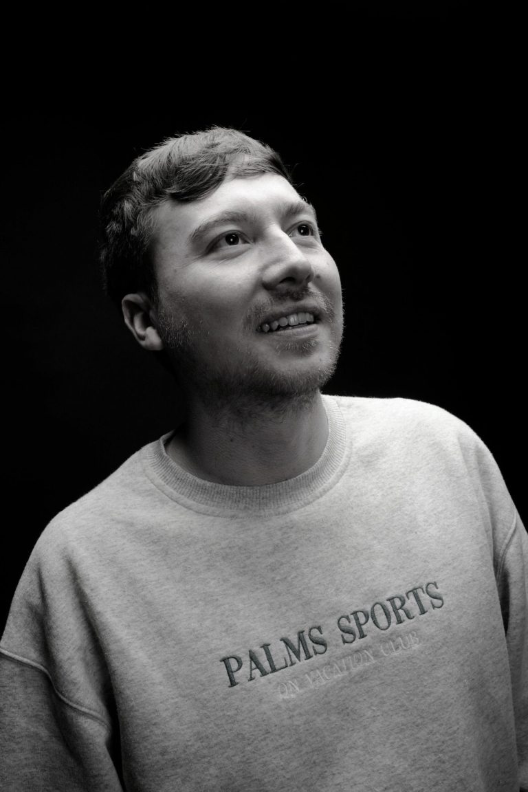 Mann mit lächelndem Gesicht, trägt ein graues Sweatshirt mit Aufdruck.