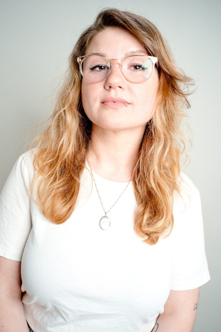 Junge Frau mit langen, lockigen Haaren und Brille, lächelt in die Kamera.