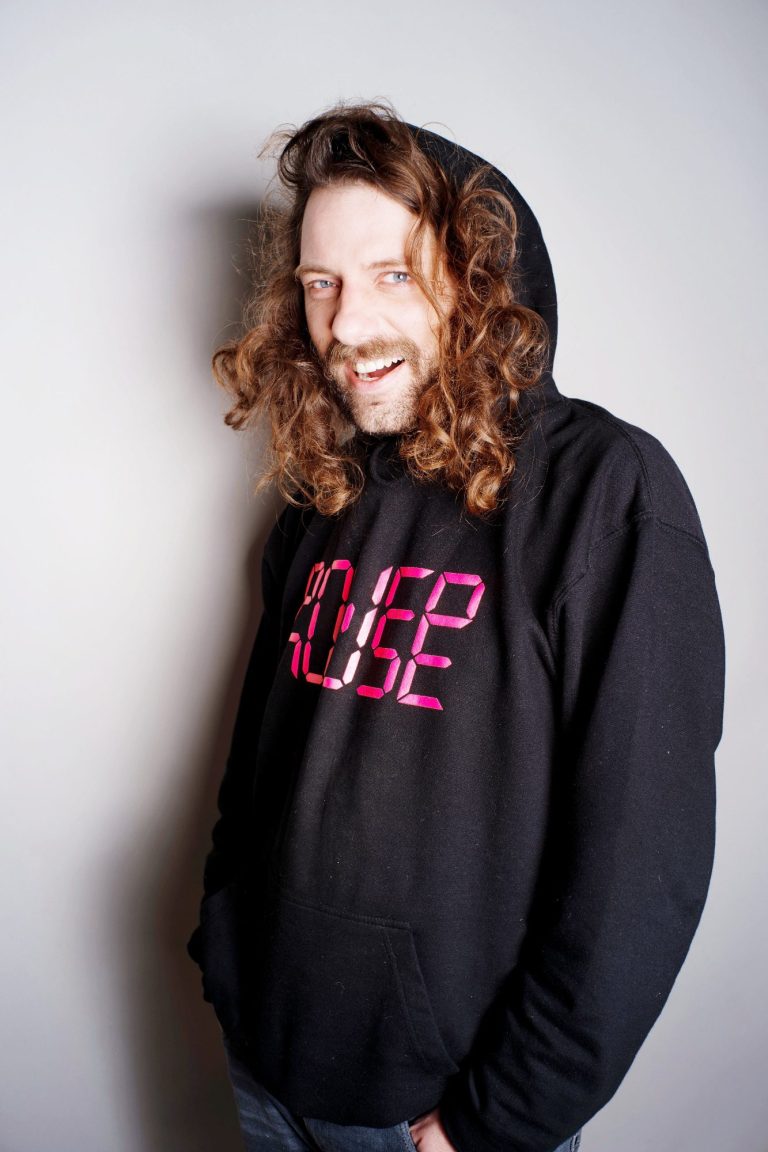 Lächelnder Mann mit lockigem Haar in einem schwarzen Hoodie mit pinkem Text.