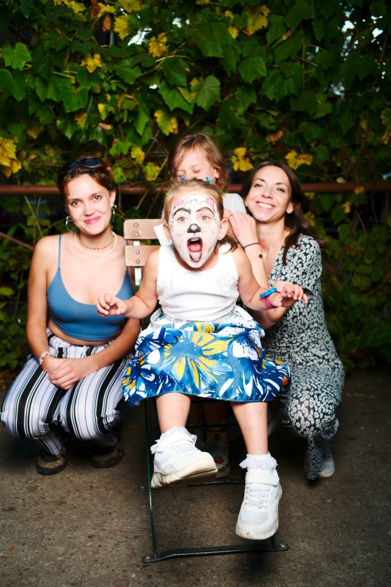 Ein Kind mit Clowns-Make-up sitzt vor zwei Frauen in bunten Outfits, grinst auffällig.
