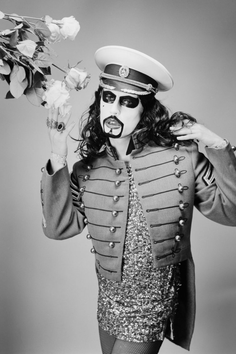 Eine Person in Militärjacke mit Sonnenbrille und Blumen, in schwarz-weiß aufgenommen.