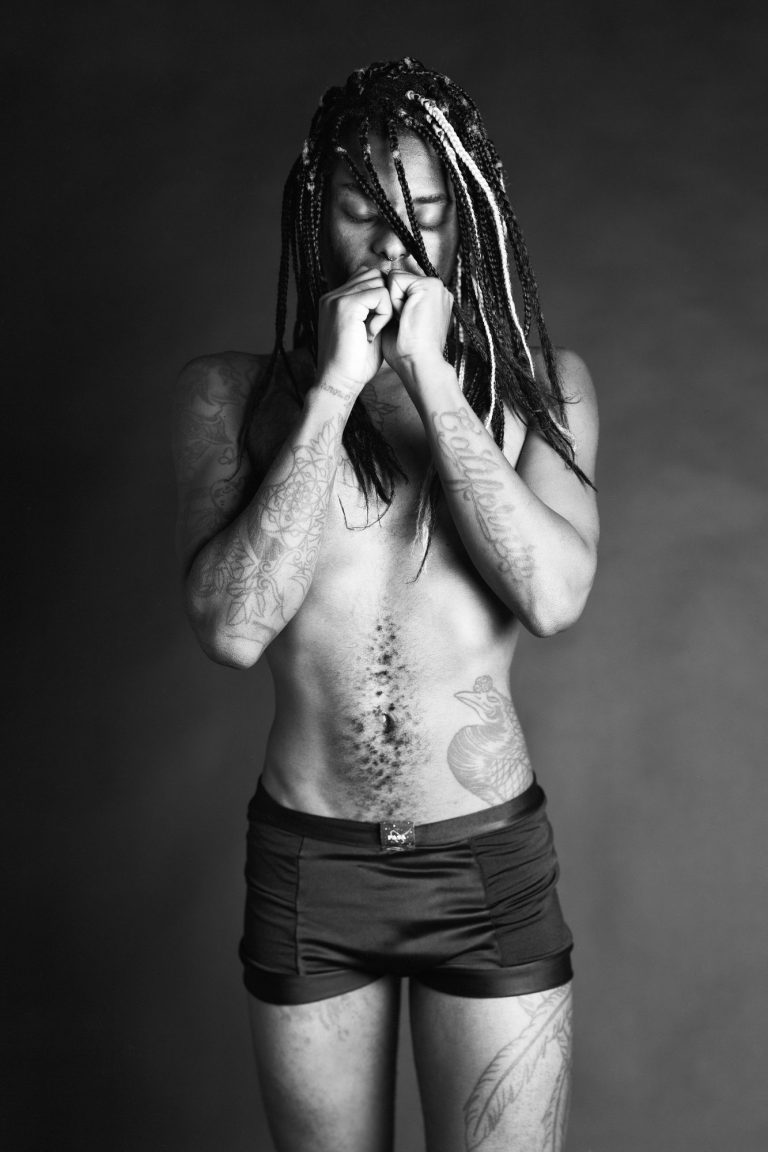 Mann mit Dreadlocks steht nackt mit verschränkten Händen vor einem neutralen Hintergrund.